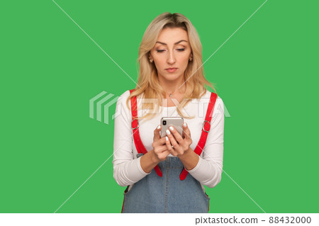 Portrait of adult pretty woman in stylish denim overalls holding smartphone and reading message, using mobile device for communication, browsing web. indoor studio shot isolated on green background 88432000