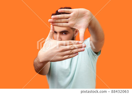 Portrait of attentive curious brunette man in casual white t-shirt looking at camera with one eye, focusing through photo frame made of fingers. indoor studio shot isolated on orange background Portrait of attentive curious brunette man in casual white t-shirt looking at camera with one eye, focusing through photo frame made of fingers. indoor studio shot isolated on orange background 88432063