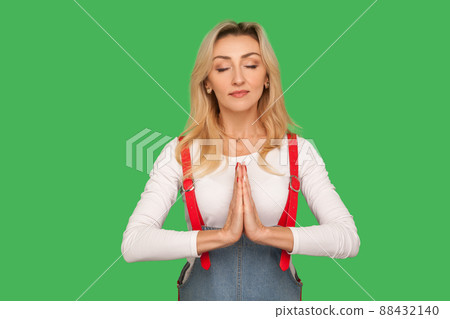 Peaceful mind, yoga practice. Portrait of beautiful calm adult woman in stylish overalls meditating with namaste gesture, feeling concentrated and relaxed. studio shot isolated on green background 88432140