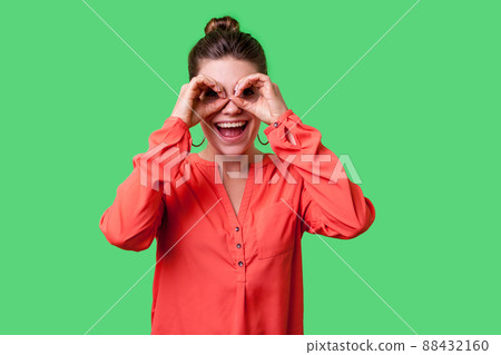 Portrait of curious lovely young woman with bun hairstyle, big earrings and in red blouse making binoculars gesture, looking far away with open mouth. indoor studio shot isolated on green background 88432160