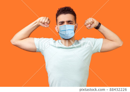 I am strong and powerful. Portrait of confident successful brunette man with surgical medical mask showing biceps, full of motivation and willpower. indoor studio shot isolated on orange background 88432226