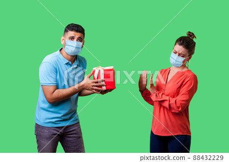 Portrait of upset resentful man with surgical medical mask giving present to naughty woman, dissatisfied girl showing refusal gesture, bad gift. isolated on green background, indoor studio shot Portrait of upset resentful man with surgical medical mask giving present to naughty woman, dissatisfied girl showing refusal gesture, bad gift. isolated on green background, indoor studio shot 88432229