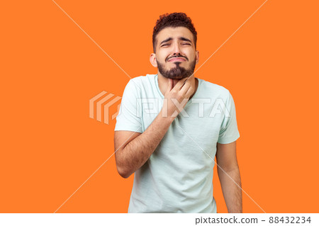 Inflamed throat. Portrait of sick brunette man with beard in white t-shirt touching painful neck, suffering sore throat, tonsillitis symptom, medical concept. studio shot isolated on orange background 88432234