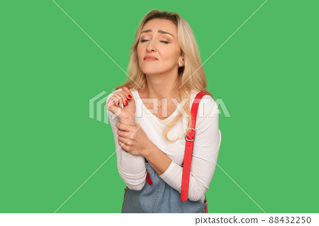 Hand injury. Portrait of adult woman in denim overalls touching her damaged arm and suffering from pain in sprained wrist, arthritis inflammation. indoor studio shot isolated on green background Hand injury. Portrait of adult woman in denim overalls touching her damaged arm and suffering from pain in sprained wrist, arthritis inflammation. indoor studio shot isolated on green background 88432250