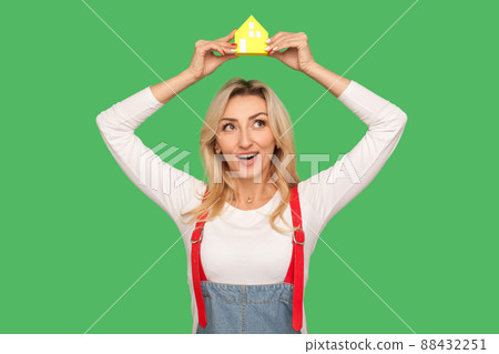 Home purchase, mortgage. Portrait of cheerful adult blond woman in urban style overalls holding paper house over head, dreaming of own accommodation. indoor studio shot isolated on green background Home purchase, mortgage. Portrait of cheerful adult blond woman in urban style overalls holding paper house over head, dreaming of own accommodation. indoor studio shot isolated on green background 88432251