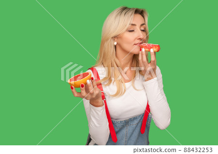 Portrait of hungry woman enjoying smell of delicious sweet donut while holding grapefruit in other hand, concept of choosing unhealthy junk food instead fruit. studio shot isolated on green background 88432253