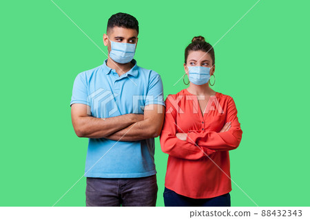 Portrait of upset couple with surgical medical mask standing together with crossed hands, looking sideways at each other with resentful glance, suspicion. isolated on green background, indoor studio Portrait of upset couple with surgical medical mask standing together with crossed hands, looking sideways at each other with resentful glance, suspicion. isolated on green background, indoor studio 88432343