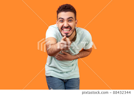 Portrait of extremely happy brunette man with beard in white t-shirt holding hand on belly, pointing at camera and laughing out loud, making fun of you. studio shot isolated on orange background 88432344