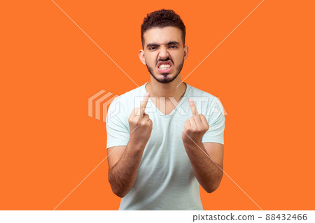Portrait of furious aggressive brunette man with beard in white t-shirt showing middle fingers, impolite rude gesture of disrespect, fuck off expression. studio shot isolated on orange background 88432466
