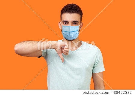 Thumbs down. Portrait of frustrated dissatisfied man with medical mask in casual white t-shirt showing dislike gesture, disapproval negative sign. indoor studio shot isolated on orange background 88432582