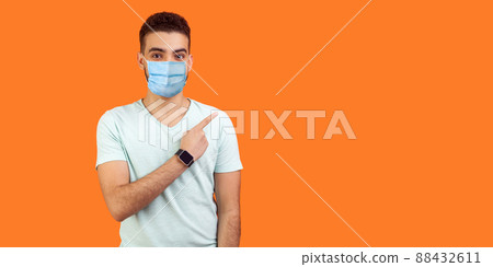 Look here. Portrait of brunette man with medical mask in casual white t-shirt standing pointing at left, empty space for text, advertise. indoor studio shot isolated on orange background Look here. Portrait of brunette man with medical mask in casual white t-shirt standing pointing at left, empty space for text, advertise. indoor studio shot isolated on orange background 88432611