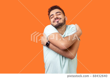 I love myself. Portrait of egoistic brunette man with beard in white t-shirt standing with closed eyes, embracing himself and smiling form pleasure and proud. indoor, isolated on orange background 88432640