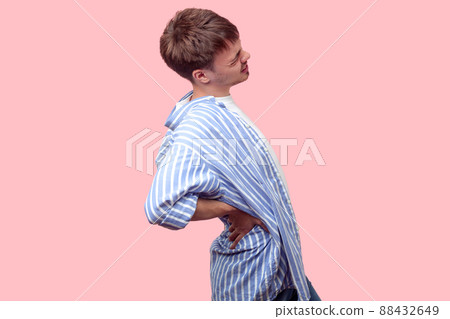 Backache. Side view portrait of young worker, sick brown-haired man in casual striped shirt with rolled-up sleeves suffering lower back pain, kidney inflammation. indoor studio shot pink background 88432649