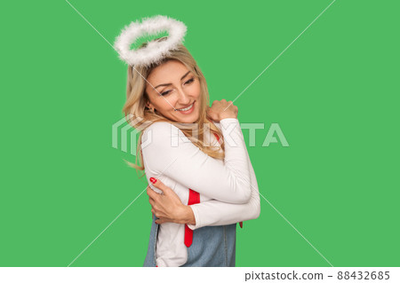 Positive self-esteem. Portrait of angelic adult woman embracing herself and smiling, satisfied with her appearance, no insecurities, self-love concept. indoor studio shot isolated on green background 88432685