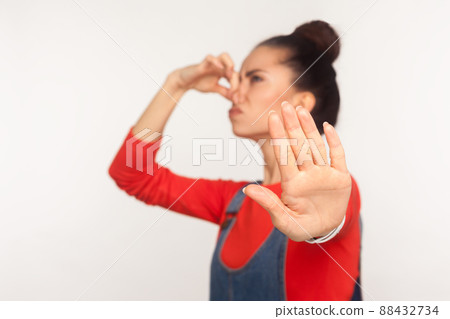 Repulsion to bad smell. Portrait of girl with hair bun in denim overalls holding breath, pinching her nose and showing stop gesture, avoiding stinky odor. studio shot isolated on white background Repulsion to bad smell. Portrait of girl with hair bun in denim overalls holding breath, pinching her nose and showing stop gesture, avoiding stinky odor. studio shot isolated on white background 88432734