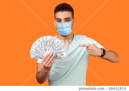 Portrait of lucky rich brunette man with surgical medical mask pointing at dollar banknotes and looking at camera with toothy smile, enjoying wealth. indoor studio shot isolated on orange background Portrait of lucky rich brunette man with surgical medical mask pointing at dollar banknotes and looking at camera with toothy smile, enjoying wealth. indoor studio shot isolated on orange background 88432814