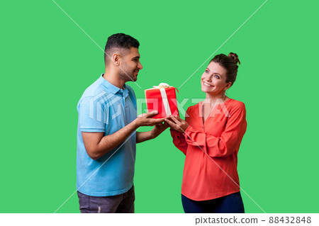 Couple celebrating Valentines day. Young handsome man in casual clothes giving box with present to beautiful elegant woman, looking at her with love. isolated on green background, indoor studio shot 88432848