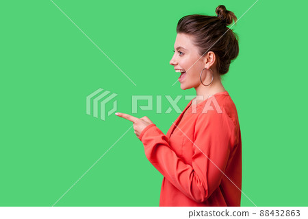Hey you. Side view portrait of amazed girl with bun hairstyle, big earrings and in red blouse pointing finger to the left, looking with astonishment. indoor studio shot isolated on green background 88432863