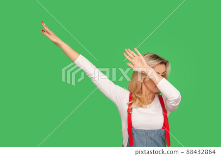Portrait of woman in stylish denim overalls raising hands in dab dance pose, celebrating success, showing gesture of triumph, famous internet move. indoor studio shot isolated on green background 88432864