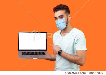 Portrait of positive joyful brunette man with surgical medical mask standing holding laptop with blank screen and smiling at camera, internet advertising. studio shot isolated on orange background 88432877