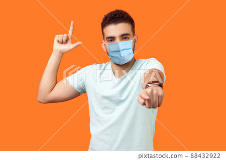 You are loser. Portrait of bossy angry man with medical mask in white t-shirt showing loser gesture and pointing at camera, mocking your defeat. indoor studio shot isolated on orange background 88432922
