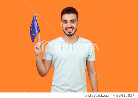 European Union flag. Portrait of happy brunette man with beard in white t-shirt smiling and holding EU flag, looking at camera with toothy smile. indoor studio shot isolated on orange background 88433034