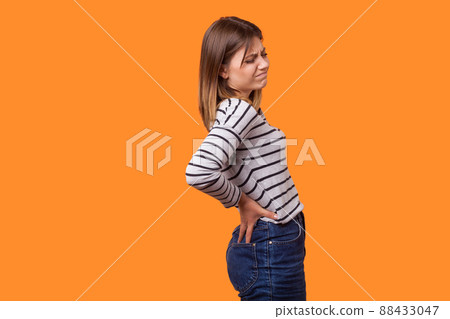 Side view portrait of sick woman with brown hair in long sleeve striped shirt standing with grimace of pain, holding back, suffering strong backache. indoor studio shot isolated on orange background 88433047