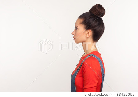 Profile of stylish slim attractive girl with hair bun in denim overalls standing with serious face, calm pensive expression, looking at side copy space. indoor studio shot isolated on white background Profile of stylish slim attractive girl with hair bun in denim overalls standing with serious face, calm pensive expression, looking at side copy space. indoor studio shot isolated on white background 88433093