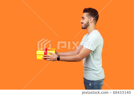 Side view portrait of positive generous brunette man with beard in white t-shirt smiling and giving gift box, sharing holiday present, charity concept. indoor studio shot isolated on orange background 88433140
