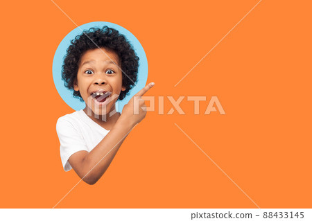 Wow look advertise here. Portrait of amazed little boy with curly hair pointing to empty place on background showing copy space for promotional ad. indoor isolated in a round hole on orange background Wow look advertise here. Portrait of amazed little boy with curly hair pointing to empty place on background showing copy space for promotional ad. indoor isolated in a round hole on orange background 88433145