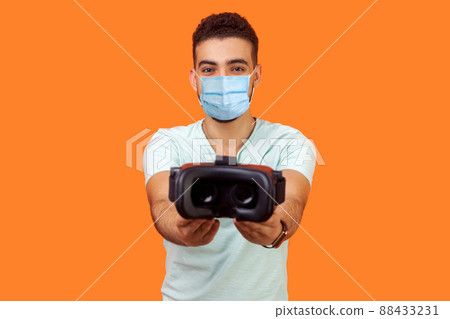 Cyberspace and innovative technologies. Portrait of cheerful man with surgical medical mask giving virtual reality headset, sharing recommending VR glasses, augmented reality. indoor studio shot 88433231