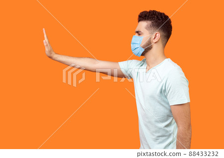 Stop, don't come any closer. Side view of serious bossy man with surgical medical mask standing holding out hand to left, prohibition gesture, saying no. studio shot isolated on orange background 88433232
