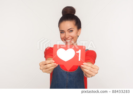 Like and follow interesting content. Portrait of charming positive trendy girl with hair bun in denim overalls showing social media heart icon to camera and smiling. indoor studio shot, isolated Like and follow interesting content. Portrait of charming positive trendy girl with hair bun in denim overalls showing social media heart icon to camera and smiling. indoor studio shot, isolated 88433283