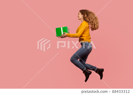 Full length portrait of cheerful excited ginger girl in sweater and denim jumping in air or flying with gift box, giving holiday present to copy space. indoor studio shot isolated on pink background Full length portrait of cheerful excited ginger girl in sweater and denim jumping in air or flying with gift box, giving holiday present to copy space. indoor studio shot isolated on pink background 88433312
