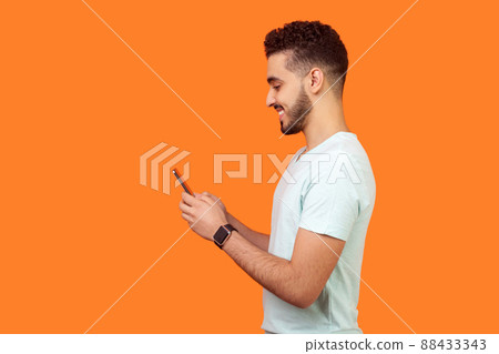 Side view of happy brunette man with beard in white t-shirt smiling while using cellphone, chatting in social media, enjoying mobile service. empty copy space for advertisement, indoor studio shot Side view of happy brunette man with beard in white t-shirt smiling while using cellphone, chatting in social media, enjoying mobile service. empty copy space for advertisement, indoor studio shot 88433343