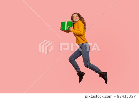 Portrait of extremely happy ginger girl in sweater and denim jumping in air with gift box, full of energy and enthusiasm flying with holiday present. indoor studio shot isolated on pink background 88433357