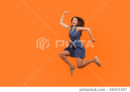 Leap to success. Full length of excited energetic motivated brunette woman in denim dress running in air, hurrying to achieve goal, jumping rejoicing victory. studio shot isolated on orange background 88433387