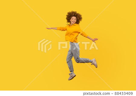 Portrait of enthusiastic lively energetic curly-haired girl in urban style hoodie and jeans flying walking in air, enjoying happy life, feeling inspired. studio shot isolated on yellow background Portrait of enthusiastic lively energetic curly-haired girl in urban style hoodie and jeans flying walking in air, enjoying happy life, feeling inspired. studio shot isolated on yellow background 88433409