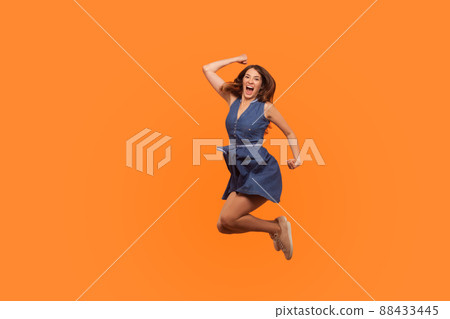 Winner joy. Full length of energetic vivid brunette woman in denim dress flying in air with inspired ecstatic expression, celebrating great success. indoor studio shot isolated on orange background 88433445