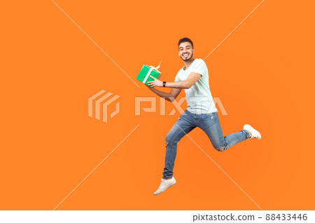 Full length portrait of excited joyous brunette man in sneakers and denim outfit smiling at camera while flying or running in air with gift box, present. studio shot isolated on orange background 88433446