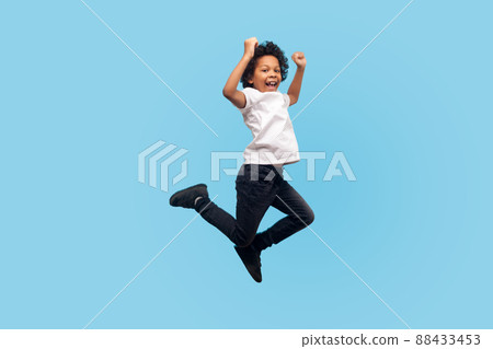 Full length, lively energetic little boy in T-shirt and denim jumping in air screaming with happiness, child flying up, feeling inspired crazy and overjoyed. indoor studio shot, blue background 88433453