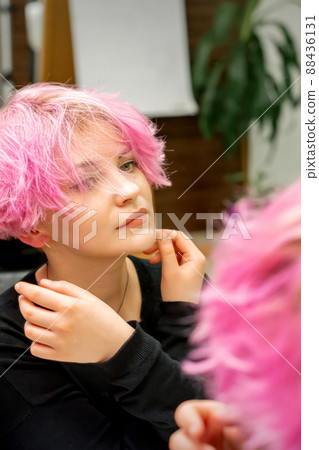 The beautiful young caucasian woman with a new short pink hairstyle looking at her reflection in the mirror checking hairstyle in a hairdresser salon 88436131