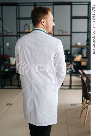 Rear view of smilng young male doctor in white medical uniform standing in hospital office. Thoughtful practitioner man posing at workplace. Concept of medical work. Rear view of smilng young male doctor in white medical uniform standing in hospital office. Thoughtful practitioner man posing at workplace. Concept of medical work. 88436795