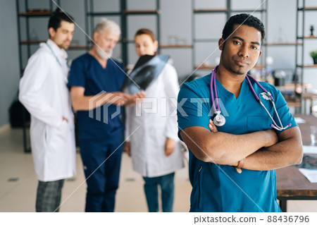 Portrait of confident African male doctor in blue uniform standing with crossed hands in medical meeting office, looking at camera. Multiethnic team of physician working on background at hospital. Portrait of confident African male doctor in blue uniform standing with crossed hands in medical meeting office, looking at camera. Multiethnic team of physician working on background at hospital. 88436796