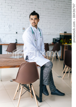 Vertical portrait of confident male doctor in white medical uniform sitting with stethoscope at table in hospital office, looking at camera. Front view of stylish practitioner man posing at workplace. Vertical portrait of confident male doctor in white medical uniform sitting with stethoscope at table in hospital office, looking at camera. Front view of stylish practitioner man posing at workplace. 88436797