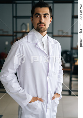 Vertical portrait of confident male doctor wearing white medical uniform standing in hospital office, looking away. Front view of bearded practitioner young man posing at workplace. Vertical portrait of confident male doctor wearing white medical uniform standing in hospital office, looking away. Front view of bearded practitioner young man posing at workplace. 88436798
