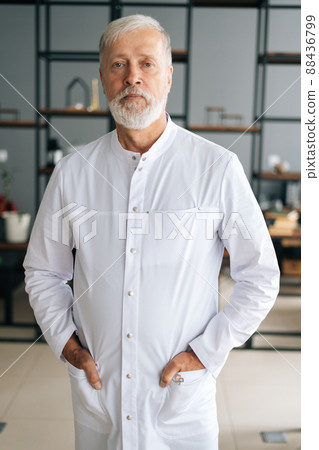 Vertical portrait of serious senior adult male doctor in white medical uniform standing in hospital office, looking at camera. Front view of gray-haired aged practitioner posing at workplace. 88436799