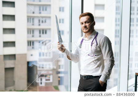 Portrait of focused male doctor in white uniform examining chest x-ray lungs scan standing near window in hospital office, thinking about diagnosis. Concept of medicine and health care. Portrait of focused male doctor in white uniform examining chest x-ray lungs scan standing near window in hospital office, thinking about diagnosis. Concept of medicine and health care. 88436800