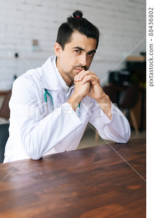 Vertical portrait of serious handsome male doctor wearing white uniform with stethoscope sitting at desk in hospital office, looking at camera. Concept of medicine and health care. 88436802