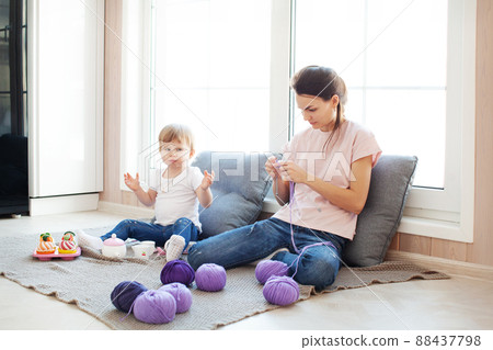 Small kid daughter play while her mother dark hair female knit, doing work or hobby in light wooden interior of house. Candid domestic life with baby 2 years, Family Small kid daughter play while her mother dark hair female knit, doing work or hobby in light wooden interior of house. Candid domestic life with baby 2 years, Family 88437798